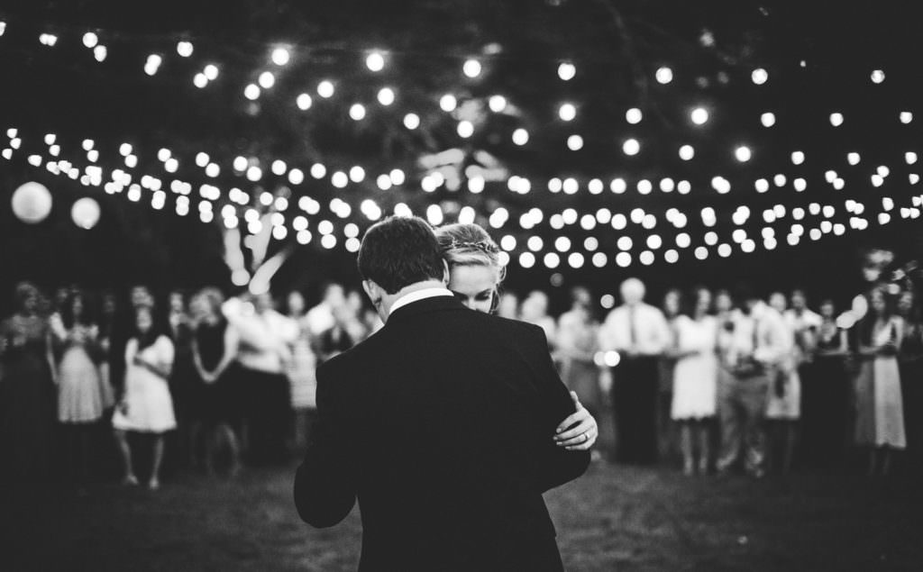 black and white photo of bride and groom's first dance with string lights and wedding guests in the background